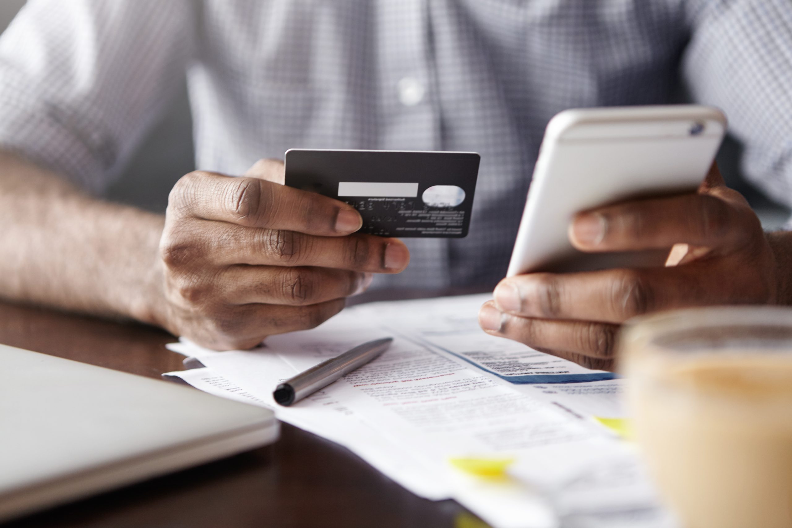 Close-up view of African man's hands holding plastic credit card and smart phone. Dark-skinned businessman using mobile banking application on his gadget while paying bills at cafe online via internet